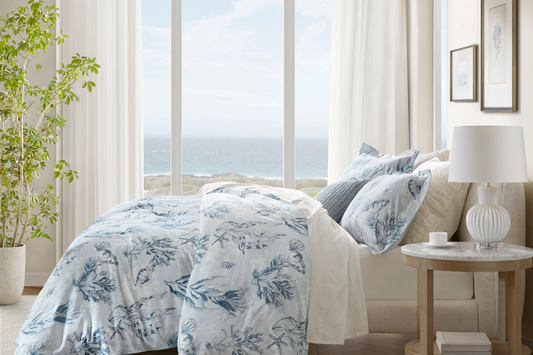 Light-filled bedroom with blue and white botanical bedding overlooking the ocean, styled with soft textures and California coastal accents.