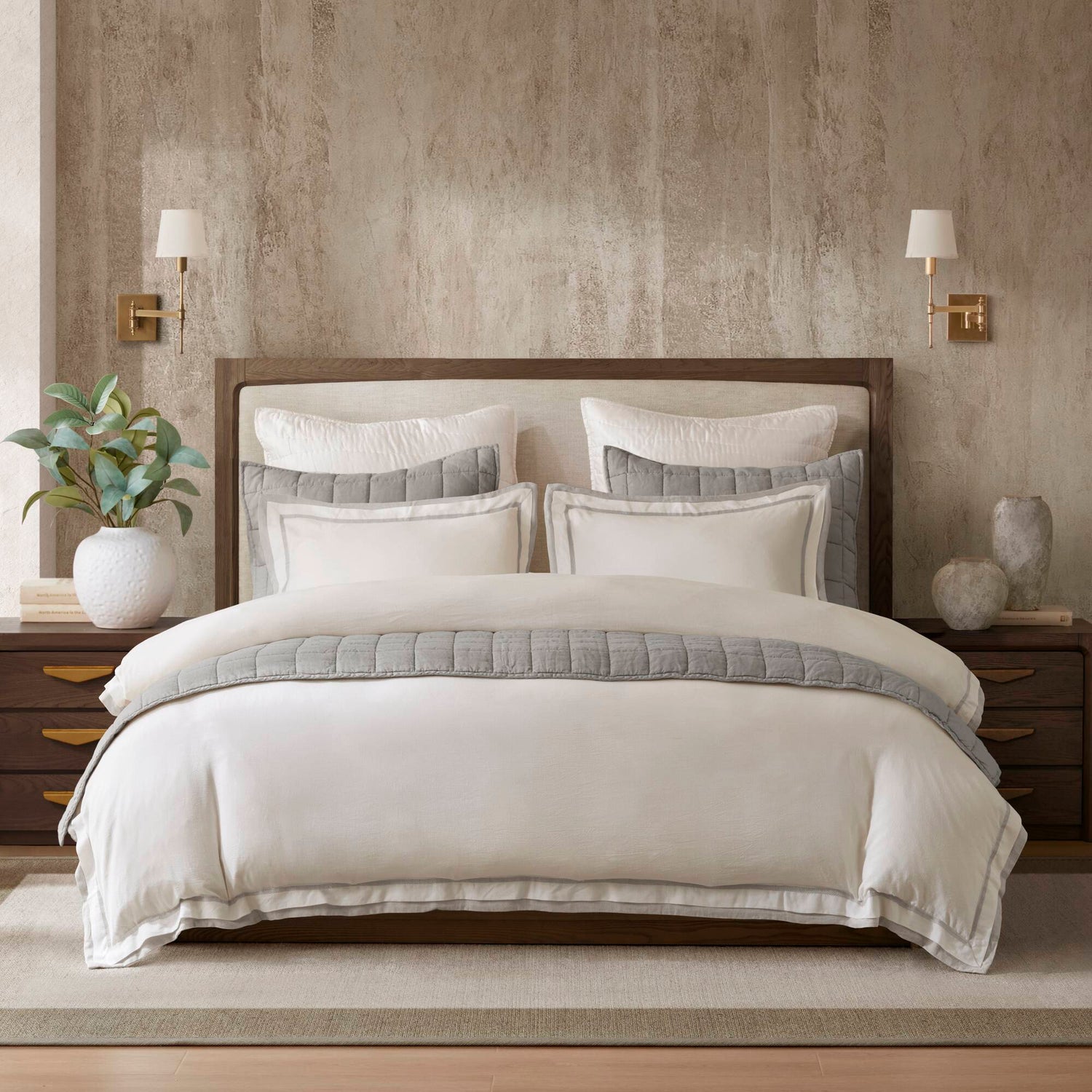 Luxurious bed with white and gray bedding, plush pillows, and a quilted comforter. There is earth-toned decor and coastal-inspired design.