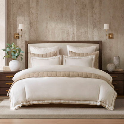 Designer bedding with beige and white tones, quilted textures, and layered pillows on a wooden bed frame, complemented by coastal-inspired decor.