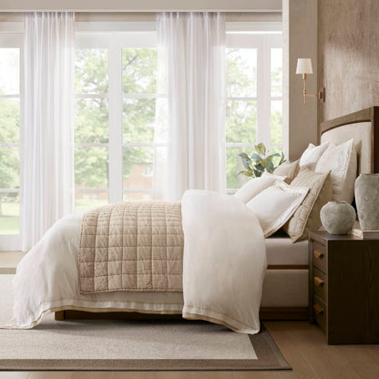 Hotel-like bed with beige quilt, white sheets, and pillows; natural light through sheer curtains; coastal-inspired decor.